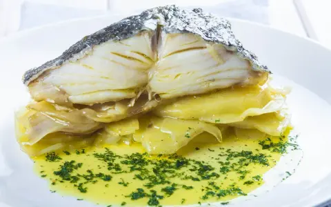 Merluza al horno con patatas, jugosa y dorada, servida sobre patatas tiernas y bañada en aceite de oliva con perejil picado.