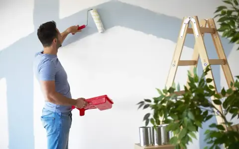 Homme peignant un mur en bleu clair, prêt à refuser assurance éco-ptz pour son projet de rénovation. Échelle et plantes à proximité.