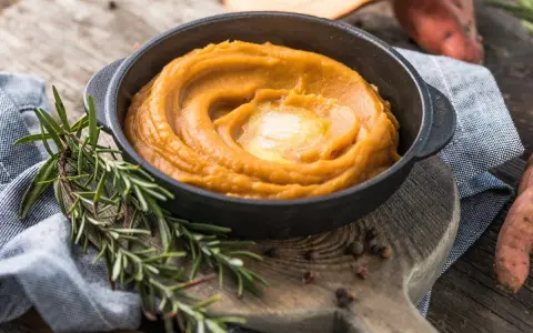 Un cremoso puré de zanahoria en un cuenco negro, coronado con mantequilla derretida. Romero fresco y granos de pimienta añaden aroma.