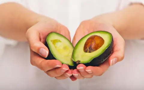Händerna håller en avokado, delad på mitten, som visar dess gröna kött och bruna kärna. En näringsrik källa till avokado vitaminer.