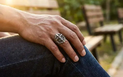 Un uomo indossa un anello con croce, simbolo di fede e protezione. Il sole illumina la sua mano poggiata sui jeans.