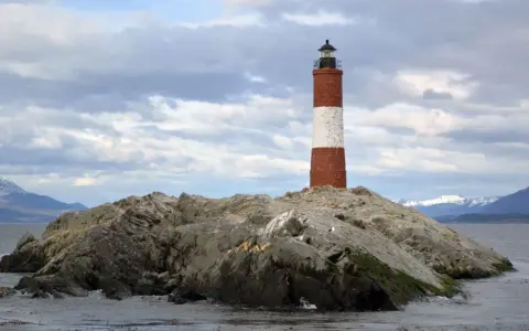 Faro a strisce rosse e bianche su un'isola rocciosa nel Canale di Beagle, con montagne innevate sullo sfondo.
