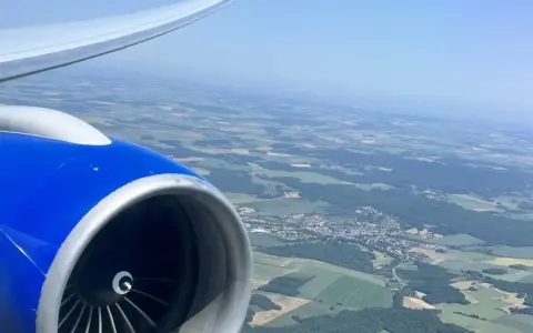 View from an airplane window, showing a blue engine and wing, with a patchwork of green fields and a small town below. Perfect for day trip flights.