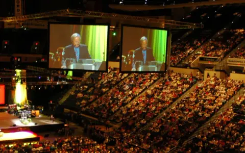 Große Menschenmenge in einer Arena, die einer Predigt von der **United Church of Christ** auf einer Leinwand folgt.