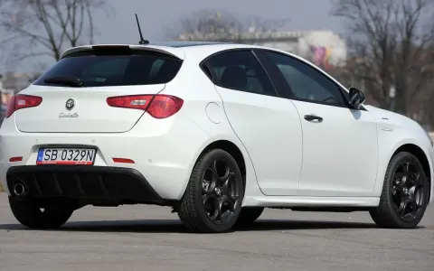 Biała Alfa Romeo Giulietta z czarnymi felgami stoi na asfalcie. Widoczny tył samochodu z tablicą rejestracyjną SB 0329N.