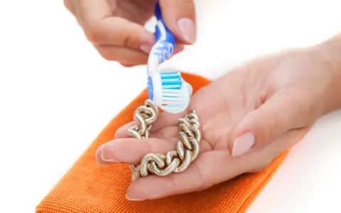 Manos limpian una pulsera de oro con un cepillo de dientes y un paño naranja. Aprende como limpiar el oro para que brille.