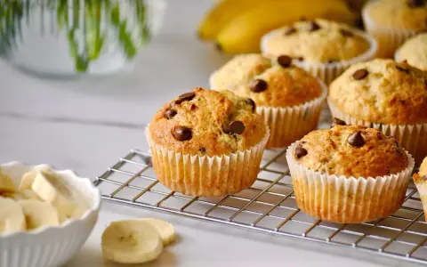 Frisch gebackene bananen muffins mit Schokostückchen auf einem Gitter, daneben eine Schüssel mit Bananenscheiben.