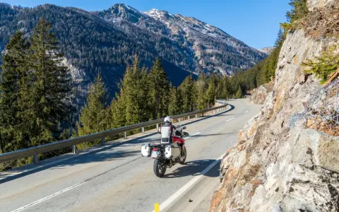 Motociclista su strada alpina in Svizzera, con montagne innevate e foreste. Ottimi consigli per un viaggio in moto in Svizzera.