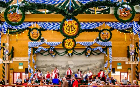 Wnętrze namiotu na Oktoberfest ozdobione niebiesko-białymi girlandami i wiankami z precelkami. Ludzie siedzą przy stołach, a w tle widać orkiestrę.