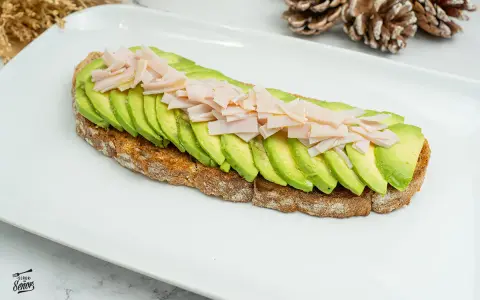Tostada integral con aguacate en rodajas y jamón picado sobre plato blanco.