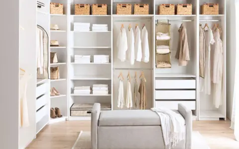 A bright, organized closet with white DIY closet shelves, featuring neatly folded clothes, baskets, and hanging garments. A grey bench sits in front.