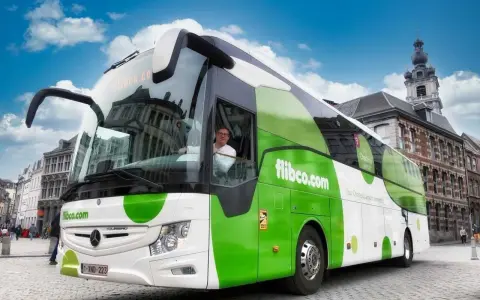 Autobus flibco.com z kierowcą, na tle miejskiej zabudowy i błękitnego nieba.