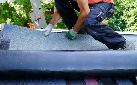Handwerker verlegt Dachschindeln. Eine Rolle Bitumenbahn liegt bereit, während die neue Schicht auf dem Dach ausgebreitet wird.