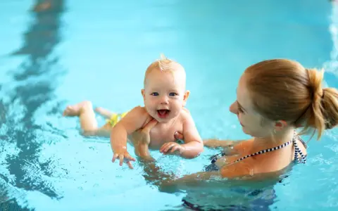 Glückliches 8 Wochen altes Baby mit Mutter im Schwimmbad. Das Baby lacht und planscht im Wasser, während die Mutter es festhält.
