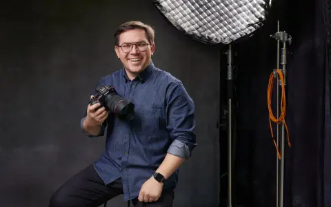 Uśmiechnięty mężczyzna z aparatem, gotowy poprowadzić kurs fotografii dla początkujących. W tle sprzęt studyjny.