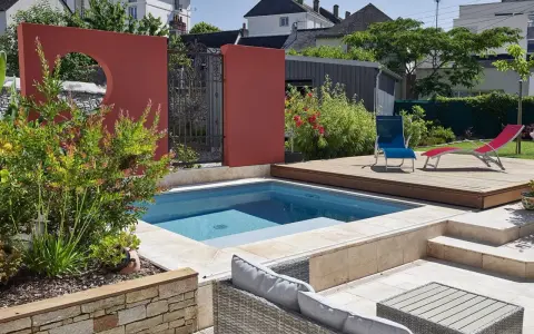 Une piscine moderne avec une belle décoration autour piscine : mur rouge, terrasse en bois, mobilier de jardin et végétation luxuriante.