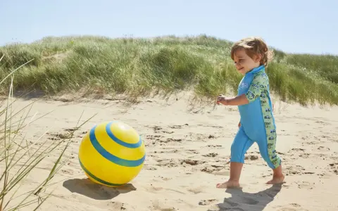 Chłopiec w niebieskim stroju kąpielowym z neoprenu bawi się żółtą piłką na plaży.