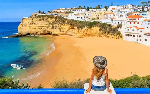 A woman in a sun hat enjoys the view of a golden beach and turquoise water in Portugal's Algarve region.