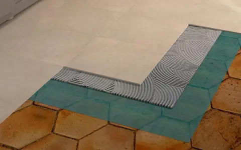Dettaglio di un pavimento in fase di posa: nuove piastrelle beige sopra uno strato di colla, membrana impermeabilizzante turchese e vecchie piastrelle esagonali color terra.