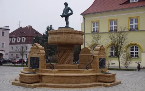 Kamienna fontanna z rzeźbą rybaka na szczycie, ozdobiona płaskorzeźbami statków.
