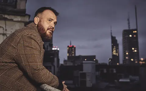 Chmielarz długa noc. Mężczyzna w płaszczu w kratę spogląda na nocne miasto z dachu.