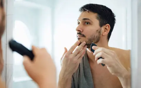 Hombre con barba corta se afeita la barba con una recortadora, buscando un afeitado apurado frente al espejo.