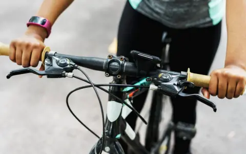 Ręce na kierownicy roweru, gotowe do jazdy. Widać manetki, hamulce i uchwyt na telefon. Trwa montaż licznika rowerowego.