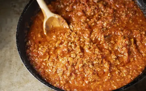 Un saporito ragù di carne in una padella, pronto per essere servito. Un cucchiaio di legno mescola la salsa ricca e densa.