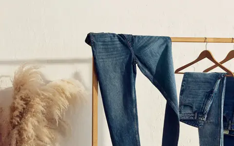 Varios pares de pantalones de **tejido denim** cuelgan de una percha de madera. A la izquierda, un gran ramo de hierba de la pampa añade un toque natural.