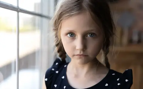 Dziewczynka z niebieskimi oczami i warkoczami, w granatowej sukience w serduszka, patrzy wprost. Jej wyraz twarzy może sugerować cechy autyzmu atypowego.