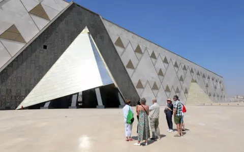 Grupa turystów podziwia nowoczesne muzeum w Gizie, którego fasada zdobiona jest hieroglifami i geometrycznymi wzorami przypominającymi piramidy.