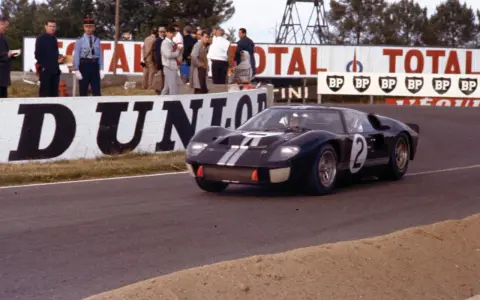 Ein schwarzer Ford GT40 Mk3 mit der Startnummer 2 rast auf der Rennstrecke, während Zuschauer und Werbetafeln im Hintergrund zu sehen sind.