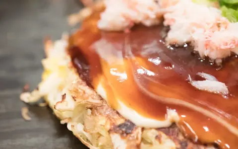 Nahaufnahme eines Okonomiyaki mit cremiger Mayonnaise und würziger Okonomiyaki-Sauce, garniert mit zarten Krabbenfleischstückchen.