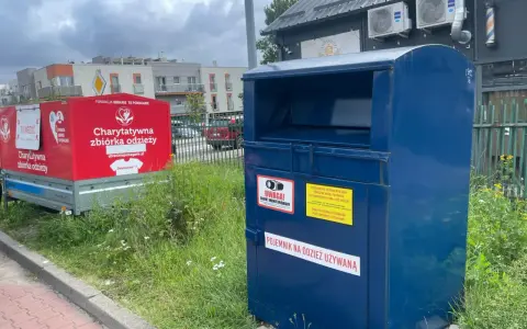 Kontener na odzież używaną obok czerwonego przyczepy z napisem "Charytatywna zbiórka odzieży". To miejsce, gdzie można oddać tekstylia, odpady ubraniowe, zamiast wyrzucać.
