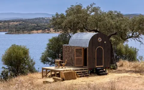 Une caravane transformée en cabane insolite, avec une terrasse en bois, au bord d'un lac paisible.