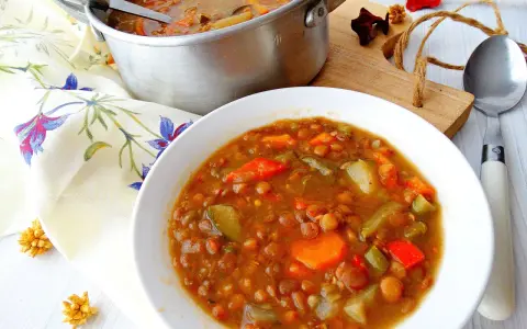 Guiso de lentejas con verduras en un cuenco blanco, junto a una olla y servilleta floral.