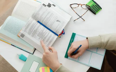 Ręce studenta nad otwartymi książkami, notatnik z napisem "I WILL BE A GREAT STUDENT", okulary i kalkulator. Nauka do egzaminu, by zdobyć tytuł zawodowy.