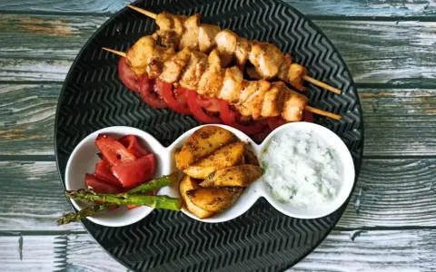 Dwa szaszłyki souvlaki z kurczaka na talerzu z pomidorami, ziemniakami, szparagami i sosem.