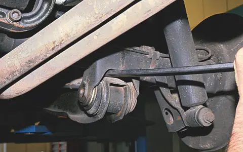 Widok z dołu na element zawieszenia samochodu. Widoczny jest metalowy wahacz z gumowymi tulejami, amortyzator i fragment układu wydechowego.