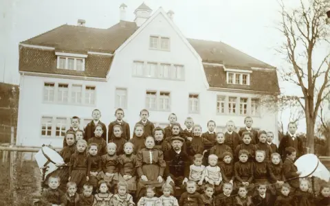 Schulkinder und Lehrer posieren vor einem Gebäude. Die Kleidung deutet auf eine Zeit hin, in der Armut, also Pauperismus, ein großes Thema war.