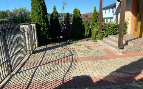 Nowy pomysł na stary chodnik: kostka brukowa w odcieniach beżu i czerwieni tworzy ciekawy wzór przed domem, otoczonym zielenią i ogrodzeniem.