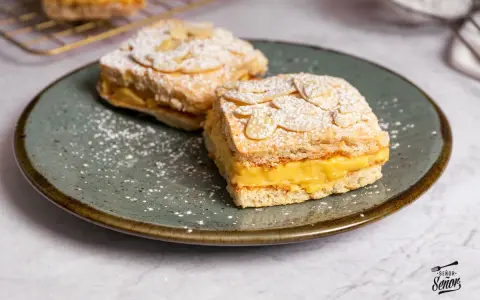 Dos porciones de pastel ruso con crema amarilla y almendras laminadas, espolvoreados con azúcar glas.
