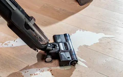 A wet/dry vacuum cleaner is cleaning up a spill of white liquid and debris on a wooden floor.