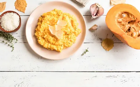 Un cremoso risotto de calabaza, decorado con láminas de queso, junto a ingredientes frescos como arroz, tomillo, ajo y una calabaza partida.