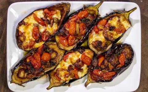 Melanzane a scarpone ripiene di pomodorini, formaggio filante e olive, cotte al forno su un piatto bianco.