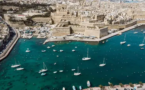 Segeln in Malta: Boote und Yachten im türkisfarbenen Wasser vor einer historischen Stadtkulisse mit Festungsmauern.