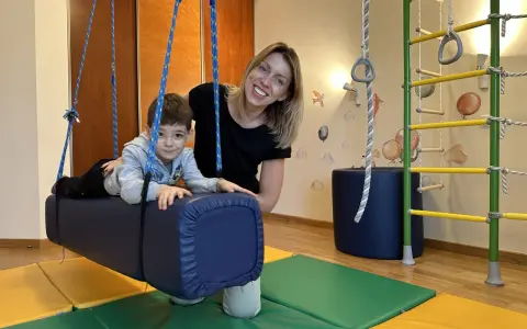 Chłopiec z ADHD u niemowlaka ćwiczy na huśtawce terapeutycznej z uśmiechniętą fizjoterapeutką.