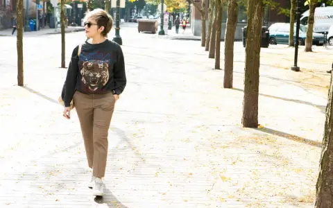 A woman walks down a tree-lined street in a trendy plaid pants outfit, featuring a graphic sweatshirt and white sneakers.