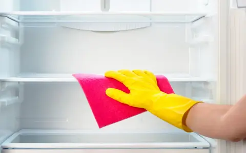 Mano con guante amarillo limpiando estante de frigorífico vacío con paño rosa.
