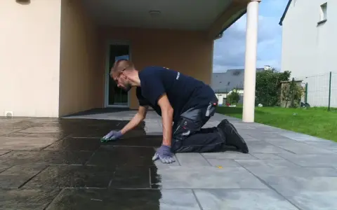 Un homme agenouillé effectue le nettoyage de dalles de terrasse avec une éponge verte, redonnant leur éclat à la pierre.